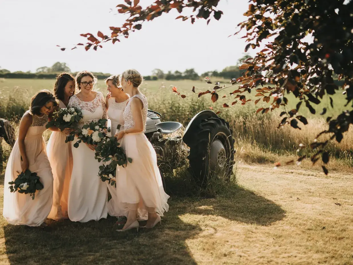 Bride and bridesmaids laughing outdoors near a rustic tractor on a sunny day.
