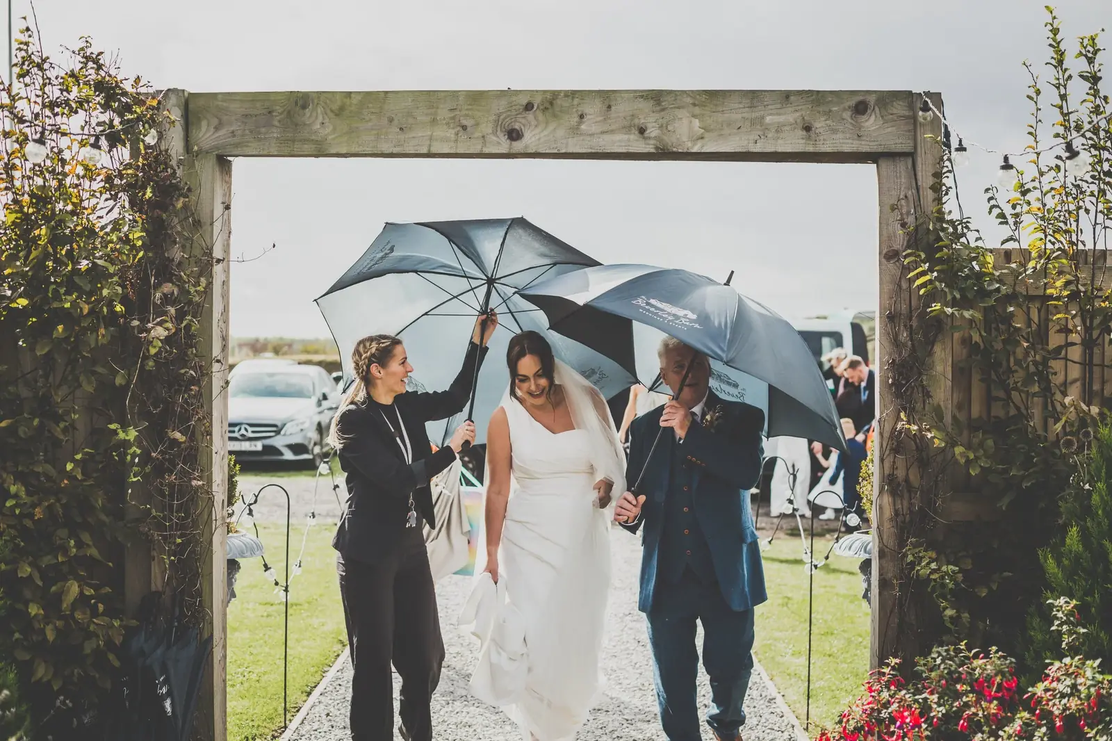 People at a wedding under umbrellas, walking through a wooden archway decorated with ivy and string lights, on a cloudy day.